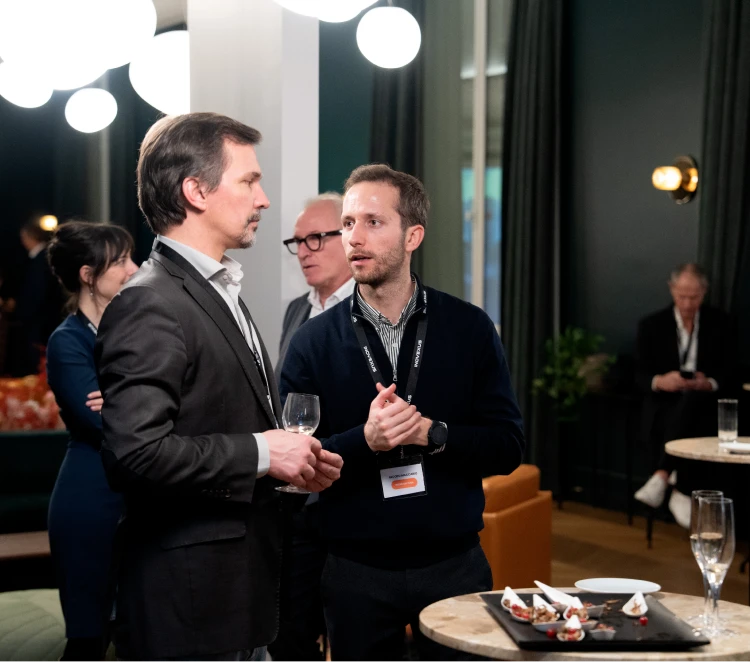 Professionals networking at a business event, engaged in conversation around cocktail tables in a modern venue, with attendees discussing opportunities and building connections.