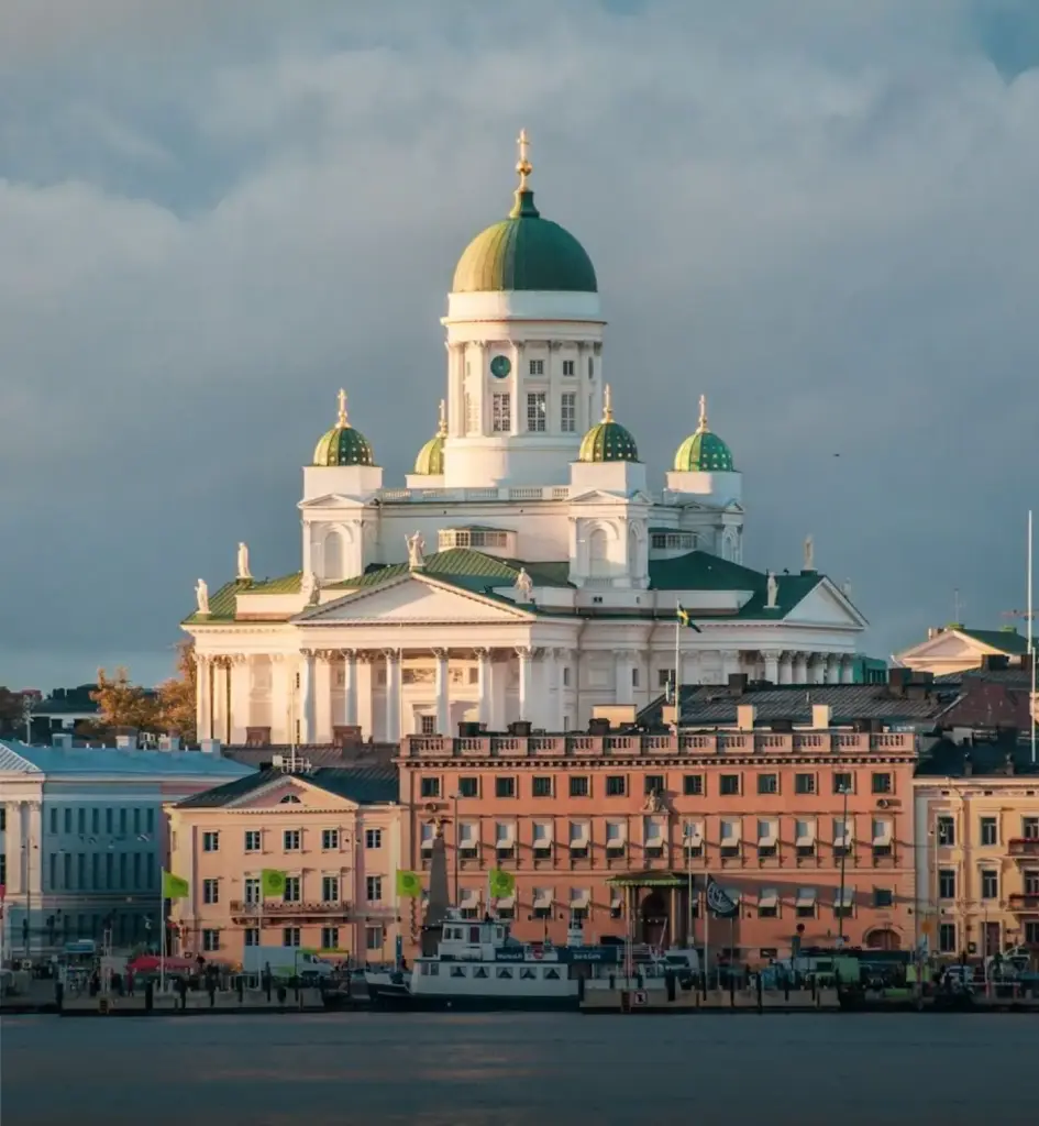 Helsinki Cathedral overlooking the city waterfront, featuring a grand white neoclassical façade, a large green central dome, and four smaller domes illuminated by warm evening light.