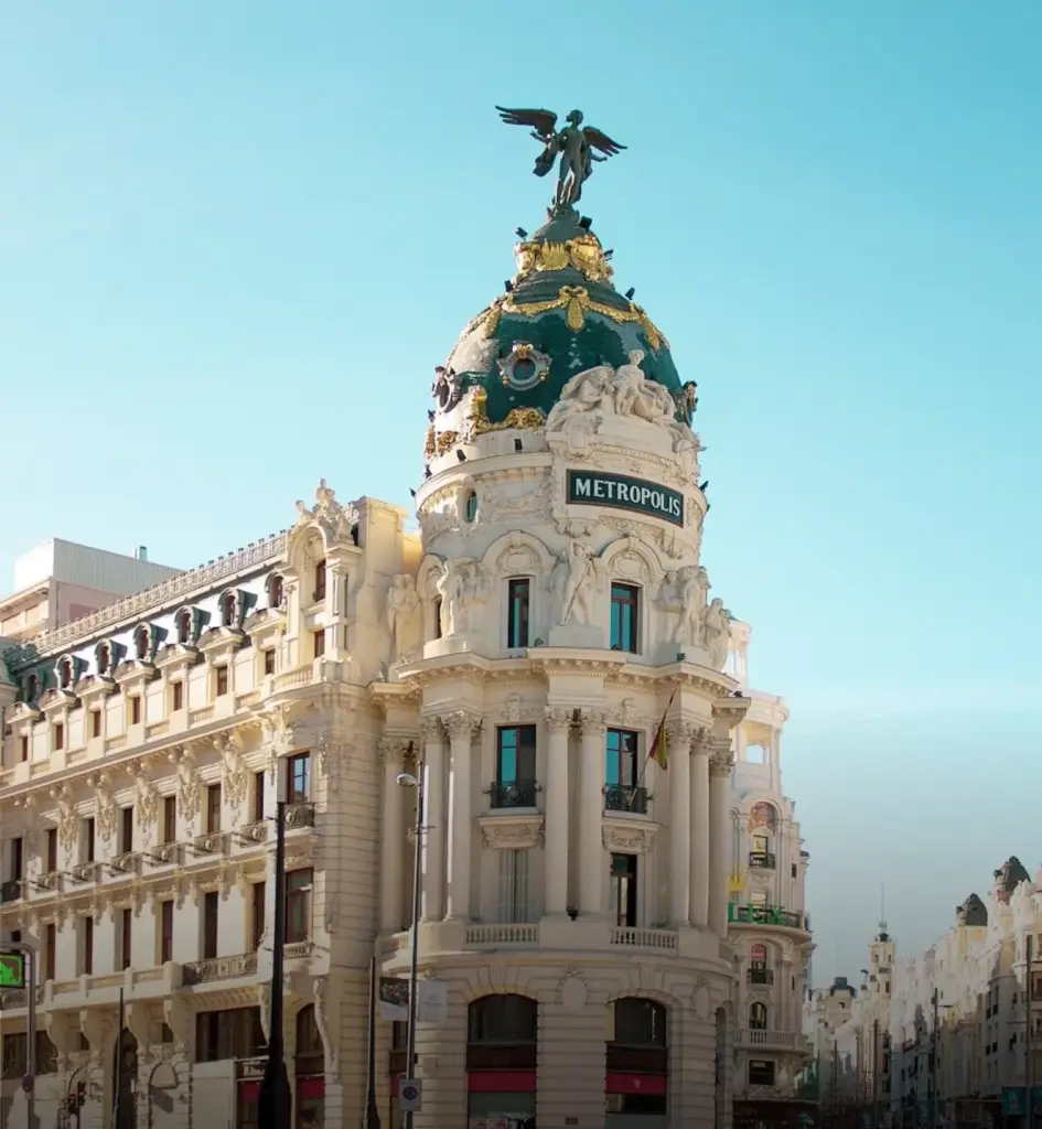 Historic Metropolis Building in Madrid, Spain, featuring an ornate white stone façade, a green-and-gold domed roof, and a winged statue at the top against a clear blue sky.