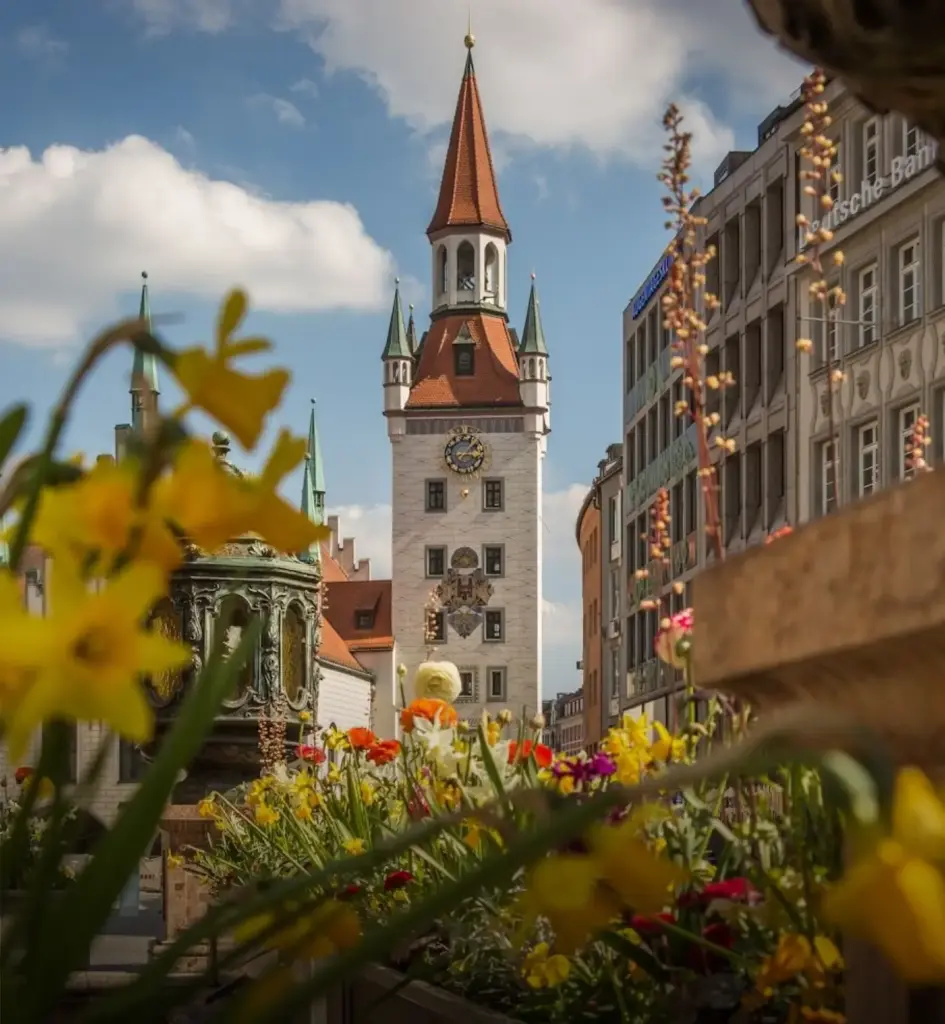 Munich’s Old Town Hall clock tower rising above a colorful flower garden, featuring a red-tiled spire, historic architecture, and a bright blue sky with scattered clouds.