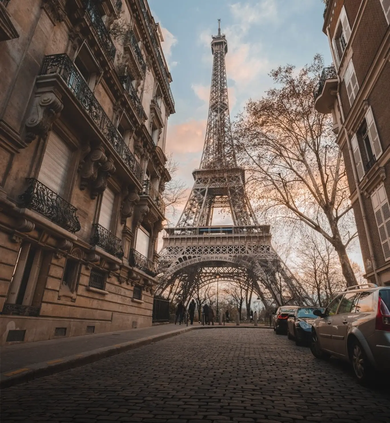 The Eiffel Tower rising above a cobblestone street in Paris, framed by historic apartment buildings, parked cars, and bare trees beneath a soft evening sky.
