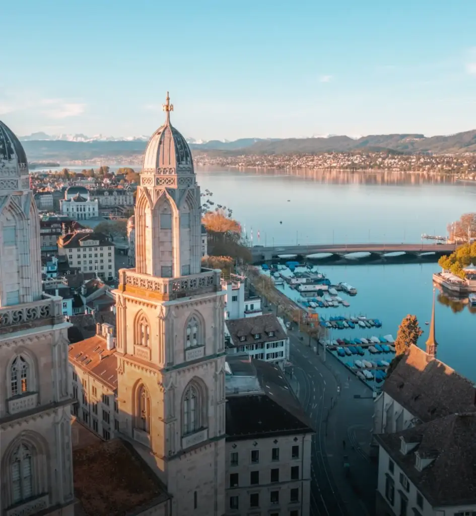 Twin towers of Grossmünster Church overlooking Zurich’s historic old town and Lake Zurich, with a bridge, marina, and distant mountains visible under a clear blue sky.
